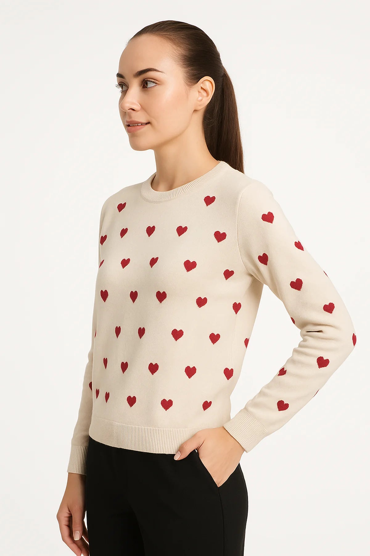 Side view of a woman wearing a natural sweater with a red heart pattern and ribbed trim, paired with black pants against a white background.