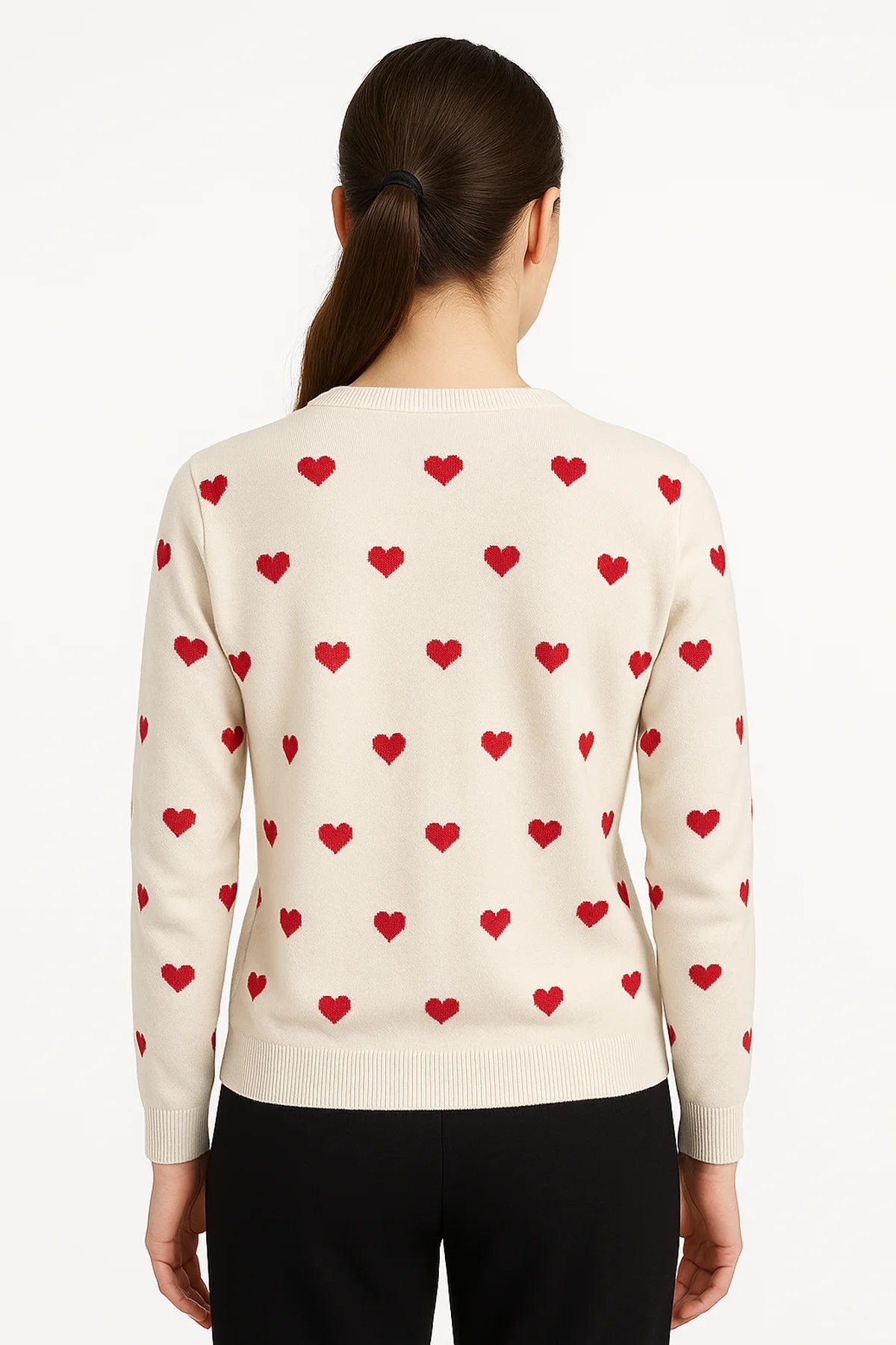 Back view of a woman wearing a natural sweater with red heart motifs and ribbed trim, styled with black pants against a white background.