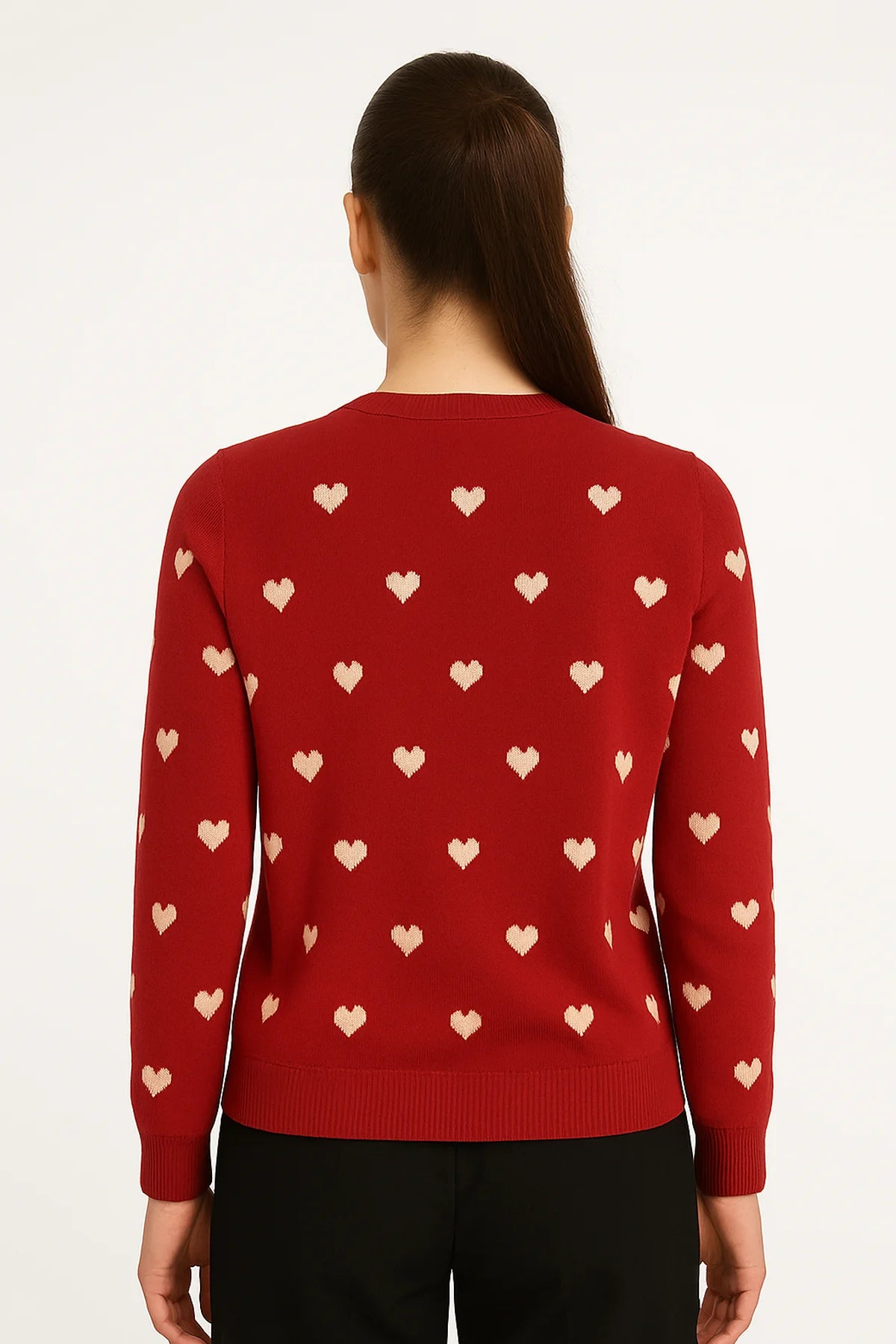 Back view of a woman wearing a red sweater with cream heart motifs and ribbed trim, paired with black pants against a white background.