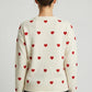 Person wearing a cream sweater with red heart patterns on a plain background